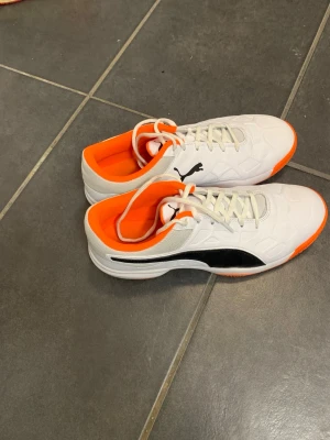 Puma vita och orange fotbollsskor - Säljer ett par Puma fotbollsskor i storlek 42.5 med snygg vit ovandel, svart Puma-logga och grymt orange innersula och yttersula. Skorna har snörning och en platt sula som passar perfekt för inomhusspel. Materialet är syntet och mesh för bra ventilation.