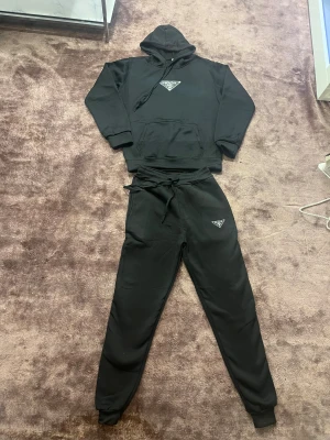 Svart hoodie från Prada med logga - Svart hoodie från Prada med klassisk logga på bröstet. Tröjan har huva med snörning, stor magficka och ribbade muddar vid ärmslut och nederkant. Materialet känns mjukt och är troligen bomull eller polyester. Perfekt för chill dagar och streetwear-stil.