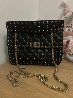 Svart quiltad axelväska med nitar - Snygg svart axelväska från Valentino i quiltat skinn med guldiga nitar och kedja. Väskan har ett vridlås i guld och en röd innerficka. Perfekt för dig som gillar edgy detaljer och lyxig känsla. Kedjan kan bäras crossbody eller på axeln. Kedjan och låsningen är lite missfärgad! 