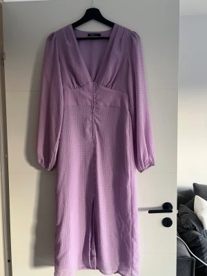 Ljuslila långklänning från Gina Tricot - Säljer en ljuslila långklänning från Gina Tricot med v-ringad hals och långa ärmar. Klänningen har ett subtilt rutigt mönster, knappar framtill och ett flowigt tyg som ger en snygg siluett. Perfekt för dig som gillar romantiska och trendiga plagg.
