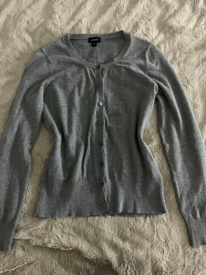 Grå kofta från Lindex  - En klassisk grå kofta från Lindex i storlek S. Koftan har rund halsringning, långa ärmar och knäppning framtill med matchande knappar. Tillverkad i mjuk bomull och passar perfekt över ett linne. 