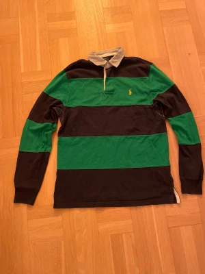 Randig rugbytröja Polo Ralph Lauren - Säljer en klassisk långärmad rugbytröja från Polo Ralph Lauren i strl S. Tröjan har breda gröna och svarta ränder, vit krage och gul logga på bröstet. Den är i  100% bomull och har en relaxed rugbykänsla. Passformen är ganska stor. Hör gärna av er om ni har några frågor! MVH Tom