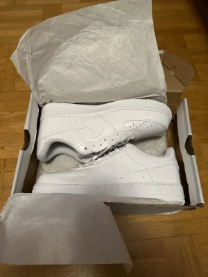 Nike Air Force 1 helvita sneakers - Nike Air Force 1 sneakers i helvit färg med klassisk låg siluett. Skorna har snörning, platt sula och rund tå. Tillverkade i skinn med perforerade detaljer på tån för extra ventilation. Snygg och tidlös design som passar till streetwear och casual outfits.