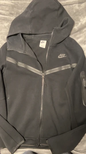 Svart Nike hoodie med dragkedja - Svart hoodie från Nike med hel dragkedja och huva. Snygga gråa detaljer över bröstet och en ficka med dragkedja på ärmen. Mjukt material som känns skönt mot huden. Nike-logga i grått på bröstet. Perfekt för chill eller träning.