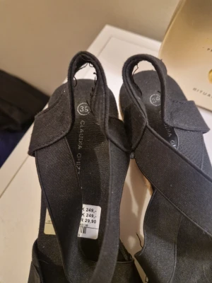 Svarta kilklack sandaler Claudia Ghizzani - Snygga svarta sandaler med kilklack från Claudia Ghizzani. Skorna har korsade band över foten och öppen häl. Ovandelen är i textil och klacken är täckt med flätad jute för en somrig vibe. Perfekta till jeans eller klänning när du vill ha lite höjd.