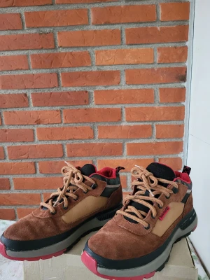 Timberland sneakers i mocka och camo - Snygga Timberland sneakers i brunt och beige mocka med svarta och röda detaljer. Skorna har grov grå sula, röd insida och coolt camouflagemönster baktill. Klassisk snörning och robust design som sticker ut.