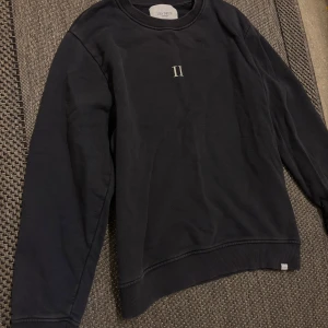 Svart sweatshirt från Les Deux - Svart sweatshirt från Les Deux med stilren vit II-logga på bröstet. Tröjan har rund hals, ribbade muddar och är tillverkad i mjuk bomull. Perfekt för dig som gillar minimalistisk design och vill ha en enkel men snygg tröja.