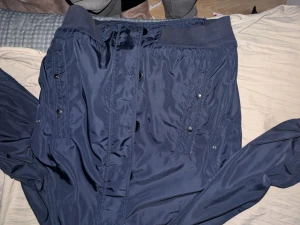 Mörkblå Moncler vindjacka med huva - Snygg mörkblå vindjacka från Moncler med huva och ribbade muddar. Jackan har flera fickor med tryckknappar, dragkedja framtill och klassisk Moncler-logga på ärmen. Perfekt för dig som vill ha en stilren och sportig look. (Äkta kvitto finns)