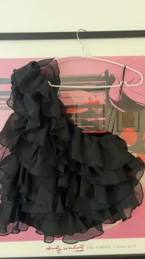 Svart volangtröja med omlott - Säljer en svart volangblus med flera lager volanger och omlott-design. Blusen har en asymmetrisk axel och är tillverkad i ett lätt, transparent material som ger en cool och luftig känsla. Perfekt för dig som gillar statement-plagg med mycket rörelse. Jättefin och i bra skick.