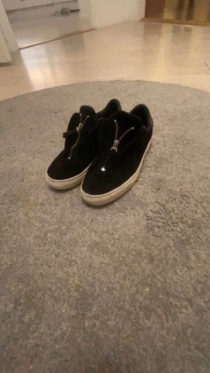 Axel arigato skor  dragkedja - Säljer ett par svarta sneakers med vit sula och snygga silverfärgade dragkedjor framtill. Skorna har rund tå och är tillverkade i mocka med detaljer i skinn. Perfekt för dig som gillar stilrena och coola skor med lite edge.