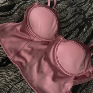 Rosa satin korsettopp från Amisu - Supersnygg rosa korsettopp från Amisu i glansigt satinliknande material. Toppen har justerbara axelband, markerade kupor och dragkedja i ryggen för enkel påklädning. Perfekt att styla med jeans eller kjol för en trendig look.