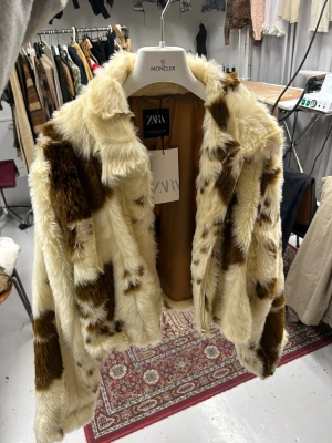 Ko pälsjacka från Zara - Säljer en fluffig pälsjacka från Zara i beige och bruna toner med ko mönster. Jackan har en kortare, loose passform, långa ärmar och är helfodrad. Perfekt för dig som vill sticka ut med en statement piece. 