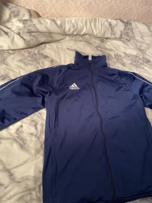 Marinblå adidas jacka med dragkedja - Säljer en marinblå adidas jacka i storlek M med vit logga på bröstet och vita detaljer längs ärmarna. Jackan har hel dragkedja framtill och är tillverkad i ett syntetmaterial som känns sportigt och bekvämt. Perfekt för träning eller chill.