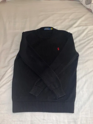 Svart stickad tröja Polo Ralph Lauren - Hej jag tänker sälja denna Svarta stickade tröja från Polo Ralph Lauren med  klassisk rund hals och röd broderad logga på bröstet. Jag är 181 och 66 kg och den passar mig perfekt! Om de är något hör av dig gärna.