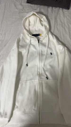 Vit hoodie från Polo Ralph Lauren - Vit hoodie från Polo Ralph Lauren med klassisk logga broderad i svart på bröstet. Tröjan har huva med snörning, dragkedja framtill och två fickor. Tillverkad i mjuk bomull och har ribbade muddar vid ärmslut och nederkant. Perfekt för chill dagar.