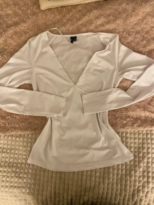 Vit långärmad v-ringad topp nelly  - Säljer en stilren vit långärmad topp från nelly med djup v-ringning. Toppen har en figurnära passform och är tillverkad i mjukt, stretchigt material som känns skönt mot huden. Perfekt att matcha med jeans eller kjol för en clean look.