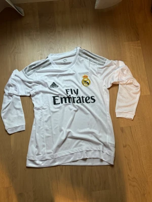 Real Madrid långärmad matchtröja Adidas - Säljer en vit långärmad Real Madrid matchtröja från Adidas med silverfärgade ränder på axlarna och klubbmärke på bröstet. Tröjan har Fly Emirates-tryck och är tillverkad i ett lätt syntetmaterial som andas. Perfekt för fotbollsfans!