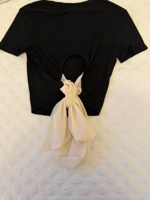 Svart croppad topp med rosett  - Säljer en svart croppad topp den  har en stor beige rosett i chiffongmaterial baktill och en öppen rygg, 