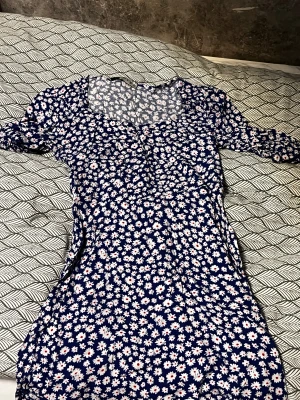 Blommig blå kortklänning - Säljer en söt blå kortklänning med vita och rosa blommor. Klänningen har en figurnära passform och korta ärmar. Perfekt för dig som gillar färgglada och lekfulla mönster. Materialet känns lätt och bekvämt mot huden.