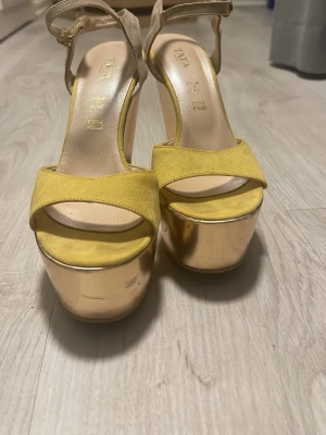 Guldiga platåsandaler med gul mocka - Säljer ett par äkta Tata Italia sandaler med höga guldfärgade platåklackar och remmar i beige mocka. Framtill har de en bred gul mockarem och öppen tå. Snygga detaljer med spänne vid ankeln och riktigt chunky plattform som ger extra höjd. De har använts mycket så skador finns undersidan på lacken alltså sulan ska tvättas se bild 6 original pris ca 500kr