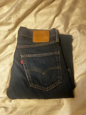 Levi's 560 mörkblå jeans W30 L32 - Mörkblå Levi's 560 jeans med klassisk femficksdesign och brunt läderpatch bak i midjan. Jeansen har raka ben och normal passform, med tydliga kontrastsömmar och röd Levi's-tag på bakfickan. Perfekta för dig som gillar tidlös denimstil.