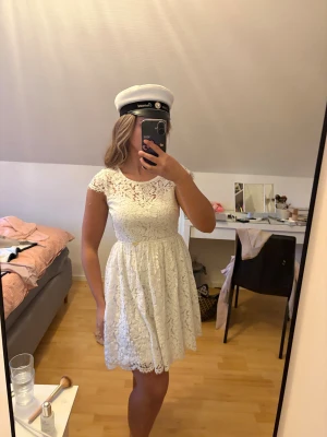 Vit spetsklänning med öppen rygg Studentklänning  - Superfin vit kortklänning i spets med blommigt mönster och rund hals. Klänningen har korta ärmar och en snygg öppen rygg med knappdetalj upptill. Perfekt till student eller sommarens alla tillfällen. Materialet är mjukt och luftigt, och midjan är markerad. Kom aldrig till användning! Helt oanvänd
