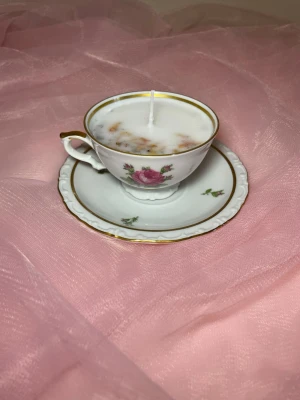 Doftljus i tekopp i en vit färg med rosa blomma, med torkade blommor nr 1 - Det här ljuset är gjort av återanvändbara material. Tekoppen är köpt secondhand och stearinet är från kyrkor som tänkte slänga det. Det här ljuset har en svag doft utav vanilj, så känsliga även kan använda det. Det finns även fler ljus med denna design på hemsidan, om man vill köpa sets. Det här ljuset har även torkade blommor på. 