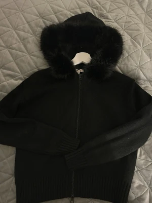 Svart stickad jacka med fuskpäls från Gina Tricot - Svart stickad jacka från Gina Tricot med huva och fluffig fuskpäls runt kanten. Jackan har dragkedja framtill och ribbade muddar vid ärmslut och nederkant. Perfekt för dig som vill ha en mysig och snygg look under kyliga dagar.