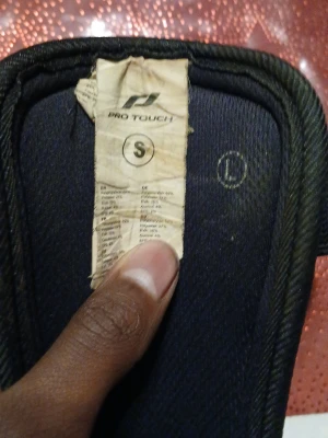 Pro Touch benskydd i neon och orange - Säljer ett par benskydd från Pro Touch i storlek S. De har en neon-gul bas med orange detaljer och svarta elastiska fästen. Ytterskalet är gjort av syntetmaterial och insidan har mjuk vaddering för extra komfort. Perfekt för dig som spelar fotboll och vill ha skydd med snygg design.