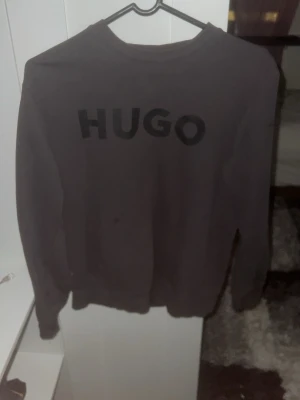 Svart sweatshirt från HUGO - Säljer en svart sweatshirt från HUGO med stor logotyp framtill. Tröjan har rund hals, ribbade muddar och lång ärm. Perfekt för dig som gillar stilrena och enkla plagg med tydlig branding.