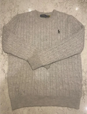 Grå kabelstickad tröja Polo Ralph Lauren - Snygg grå kabelstickad tröja från Polo Ralph Lauren i storlek M. Tröjan har rund hals, ribbade muddar och det klassiska broderade logot på bröstet. Perfekt för dig som gillar stilrena och tidlösa plagg med en exklusiv känsla.
