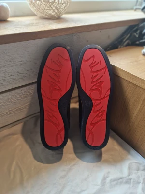 Mörkblå Christian Louboutin sneakers - Säljer ett par mörkblå sneakers från Christian Louboutin med ikoniska röda sulor och broderad logga på hälen. Skorna har rund tå, snörning och är tillverkade i mocka med snygga detaljer. Perfekt för dig som vill sticka ut med exklusiv stil.