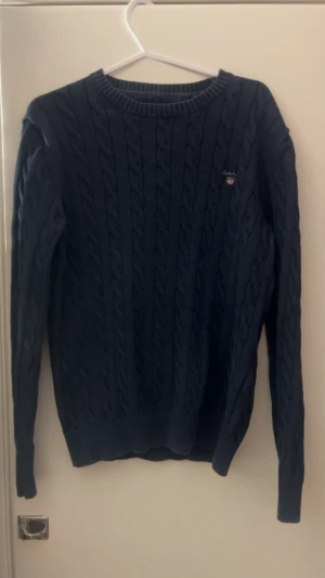 Mörkblå kabelstickad tröja från GANT - Säljer en mörkblå kabelstickad tröja från GANT i storlek M. Tröjan har rund hals, ribbade muddar och ett litet broderat GANT-emblem på bröstet. Perfekt för dig som gillar klassisk och stilren design med lite preppy känsla.