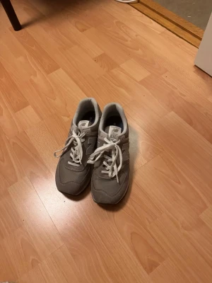 Grå New Balance 574 sneakers - Säljer ett par grå New Balance 574 sneakers med vit sula och vita snören. Skorna har en klassisk rund tå och är tillverkade i en mix av mocka och syntetmaterial. Den ikoniska N-loggan syns tydligt på sidan. Perfekt för dig som gillar stilrena och bekväma sneakers.