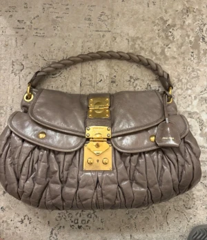 Miu Miu väska  - Säljer min sjukt fina väska från Miu Miu. Finns ett kortare band och ett längre man kan sätta på och ta av. Perfekt storlek för skolan eller om man vill ha en lite större väska. Fint skick! Använd och är lite slitet på själva metallen annars fin! Är lite grå/beige i färgen!❤️☺️