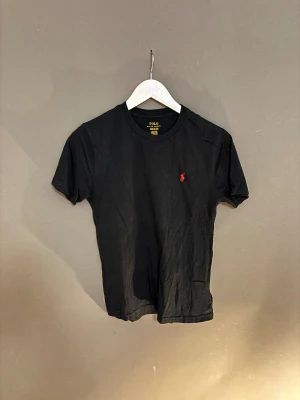 Svart t-shirt från Polo Ralph Lauren - Svart t-shirt från Polo Ralph Lauren med klassisk rund hals och röd broderad logga på bröstet. Tillverkad i mjuk bomull och har en normal passform. Perfekt för en clean och stilren look.