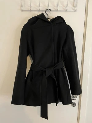 Svart kappa med huva och bälte - Snygg svart kappa med huva och knytbälte i midjan. Jackan har en stilren design med långa ärmar och är tillverkad i ett mjukt ull-liknande material. Perfekt för dig som vill ha en klassisk look med lite edge.