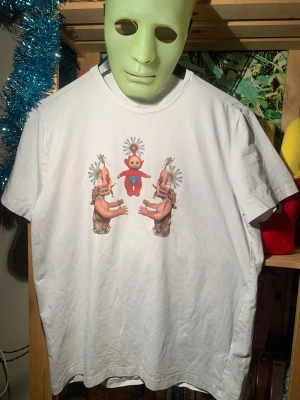 Vit t-shirt med surrealistiskt tryck - Vit t-shirt med ett unikt och surrealistiskt tryck framtill som föreställer en röd teletubby och två konstnärligt kollageartade figurer. T-shirten har klassisk passform och rund hals. Perfekt för dig som gillar statement-plagg och konstnärliga motiv.