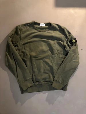 Olivgrön sweatshirt från Stone Island - Säljer en olivgrön sweatshirt från Stone Island Junior med klassisk rund hals och ribbade muddar. Tröjan har den ikoniska Stone Island-patchen på vänster ärm och är tillverkad i mjuk bomull. Perfekt för dig som gillar streetwear och snygga detaljer.
