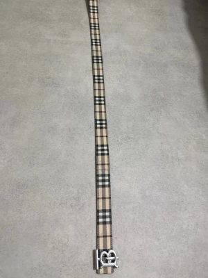 Rutig Burberry bälte med B-spänne - Snyggt Burberry bälte med klassiskt rutigt mönster i beige, svart och brunt på ena sidan och svart med präglat mönster på andra. Bältet har ett stilrent metallspänne med B-logga. Perfekt för att lyfta din outfit med en lyxig touch.