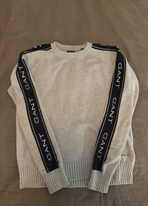 Grå stickad tröja från GANT - Snygg grå stickad tröja från GANT med marinblå band längs ärmarna där GANT-loggan är broderad i vitt. Tröjan har rund hals, ribbade muddar och nederkant. Perfekt för dig som gillar sportig och stilren design.