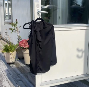 Svart bluss/festtop - Elegant svart topp/blus med asymmetrisk design och fina volanger framtill som ger ett stilfullt uttryck. Ärmlös modell med knytning vid halsen och ett luftigt, följsamt material som faller snyggt. Passar lika bra till fest som till en uppklädd vardagslook. I gott skick.