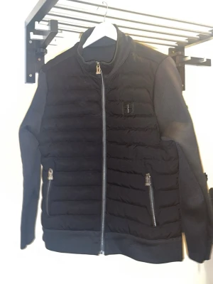 Svart pufferjacka från Stone Island - Säljer en svart pufferjacka från Stone Island med quiltad framsida och två fickor med dragkedja. Jackan har ribbade muddar, hög krage och snyggt broderad logga på bröstet. Materialet är polyester och jackan har en normal passform.