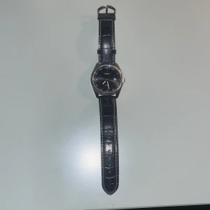 Svart Casio klocka med läderband - Stilren Casio klocka med svart urtavla och silverfärgat boett. Bandet är svart och har ett snyggt krokodilpräglat mönster i läder. Urtavlan har datumvisning och tydliga markeringar. Perfekt accessoar för dig som gillar klassisk design.