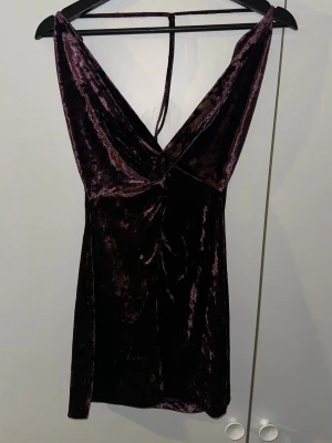 Mörklila sammetsklänning med snörning - Säljer en mörklila kortklänning i mjuk sammet med snygg snörning i ryggen och djup v-ringning. Klänningen är ärmlös och har en lyxig glansig yta som ger en cool vibe. Perfekt för dig som gillar att sticka ut med stil.