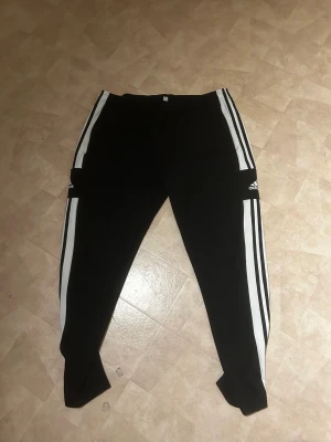 Svarta Adidas mjukisbyxor med vita ränder - Säljer ett par svarta mjukisbyxor från Adidas med klassiska vita ränder längs benen och Adidas-logga på låret. Byxorna har elastisk midja och är gjorda i mjukt polyester-material. Perfekta för chill eller träning.