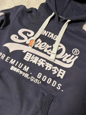 Superdry  - Säljer en superdry hoodie i marinblå färg med vit text på framsidan, det står även 54 på högerarmen, hoodien är sparsamt använd med inga defekter. 