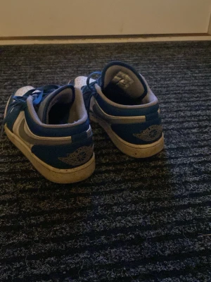 Blå Nike Jordan 1 sneakers lows - Säljer ett par blå och vita Nike Jordan 1 sneakers i low-modell med klassisk Air Jordan-logga på hälen. Skorna har snörning, rund tå och platt sula. Perfekta för dig som gillar streetwear och vill ha en ikonisk sneaker i din samling.