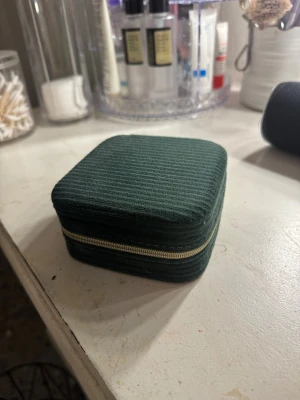 Grön smyckes necessär med guldig dragkedja - Säljer en mörkgrön smyckes necessär i manchestertyg med snygg ribbad struktur. Necessär har en praktisk dragkedja framtill i guld och en fyrkantig form som ger en cool vibe. Perfekt för dig som vill sticka ut med en unik och trendig look.
