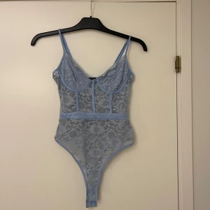 Blå spetskorsett från NAANAA - Säljer en ljusblå korsett-topp från NAANAA i spets med blommigt mönster. Toppen har tunna axelband, markerade kupor och transparent spetsmaterial. Perfekt att styla med jeans eller kjol för en trendig look.